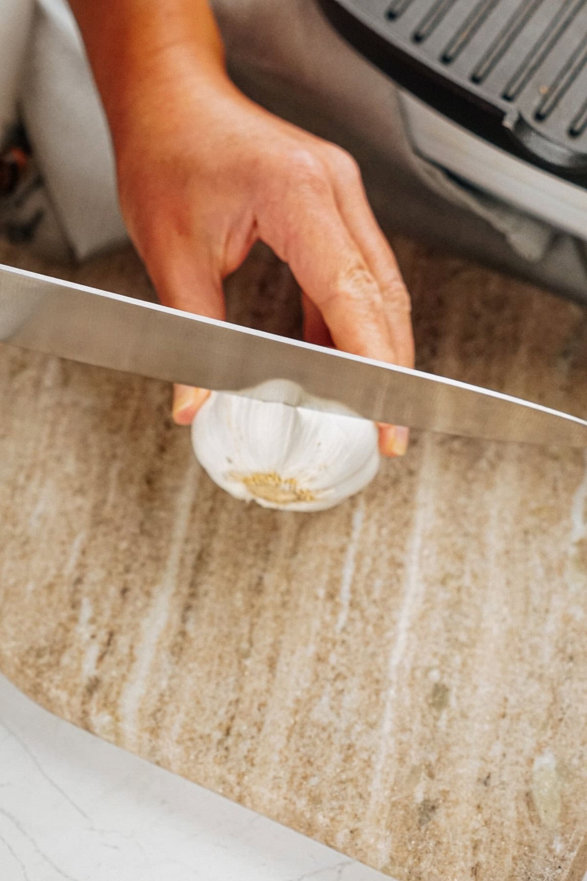 A hand holding a knife is slicing a bulb of garlic on a wooden cutting board.