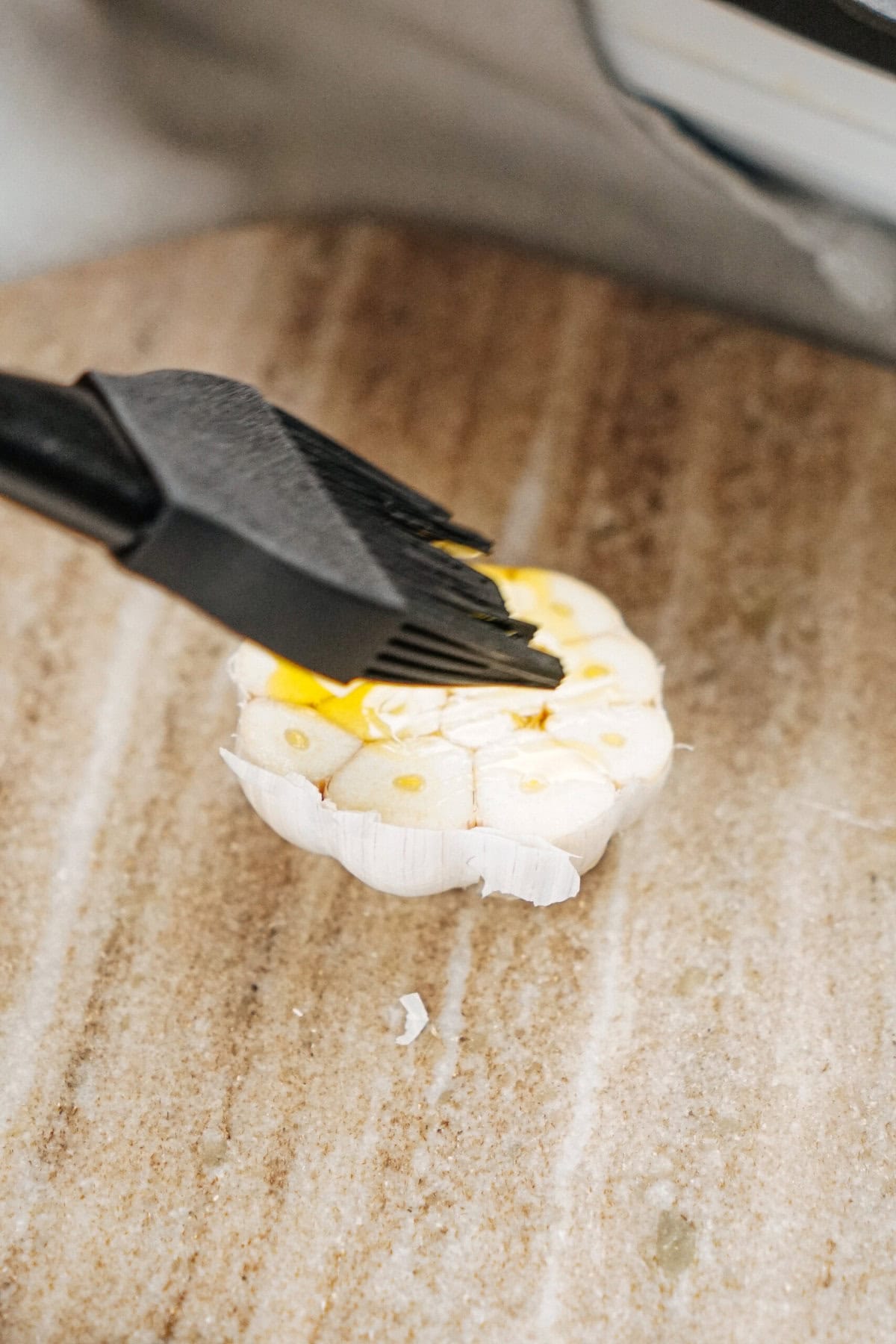 A basting brush applies oil onto a halved garlic bulb on a wooden surface.