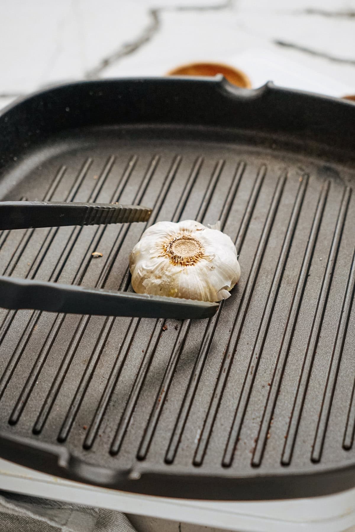 A garlic bulb is being held by tongs on a black grill pan.