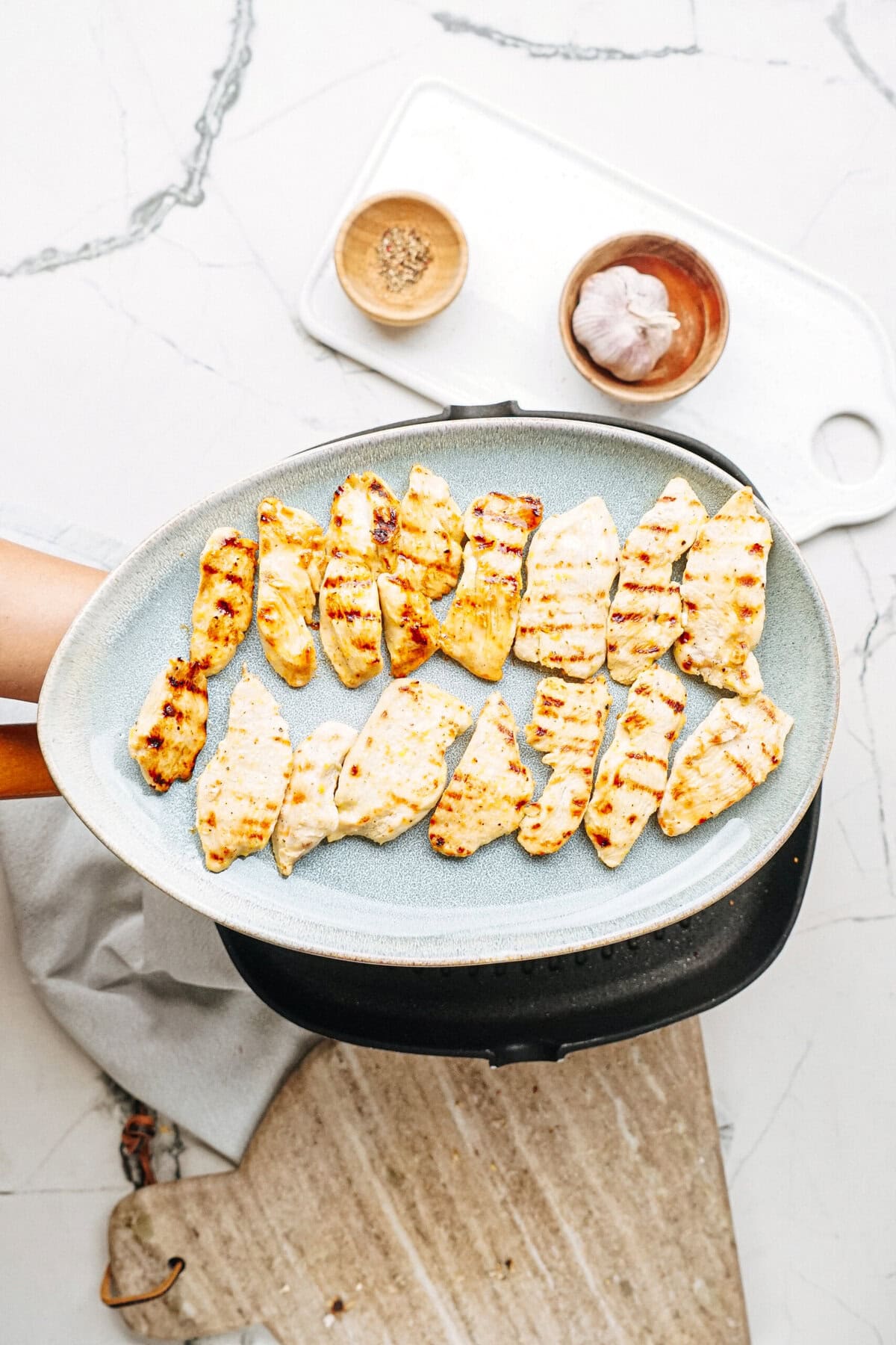 A frying pan with grilled chicken strips on a countertop, next to small bowls of spices and garlic.