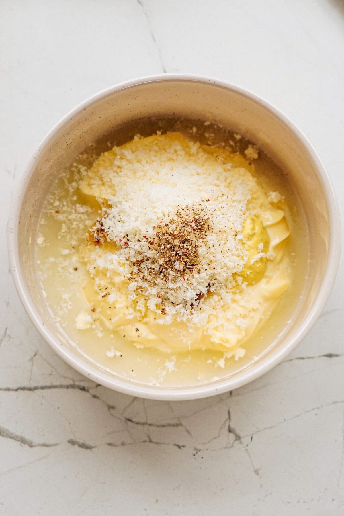 A bowl containing grated cheese, butter, and seasoning on a light marble surface.