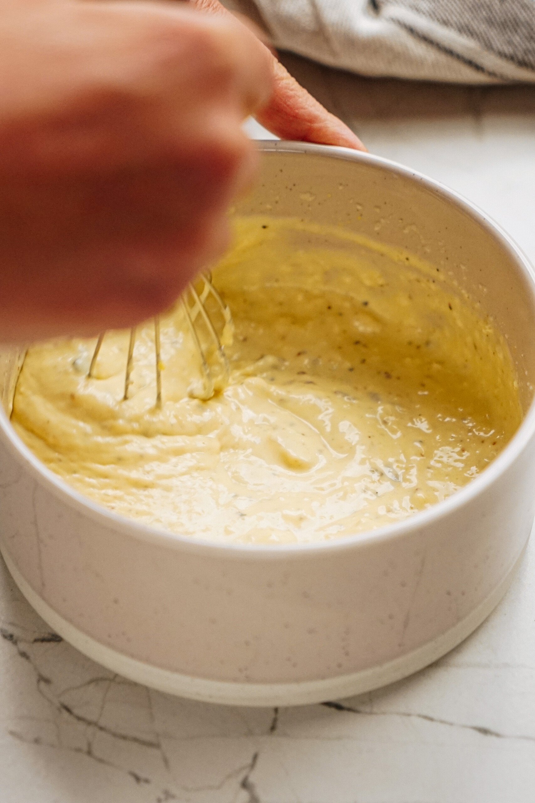 Hands whisking yellow batter in a white bowl on a marble surface.