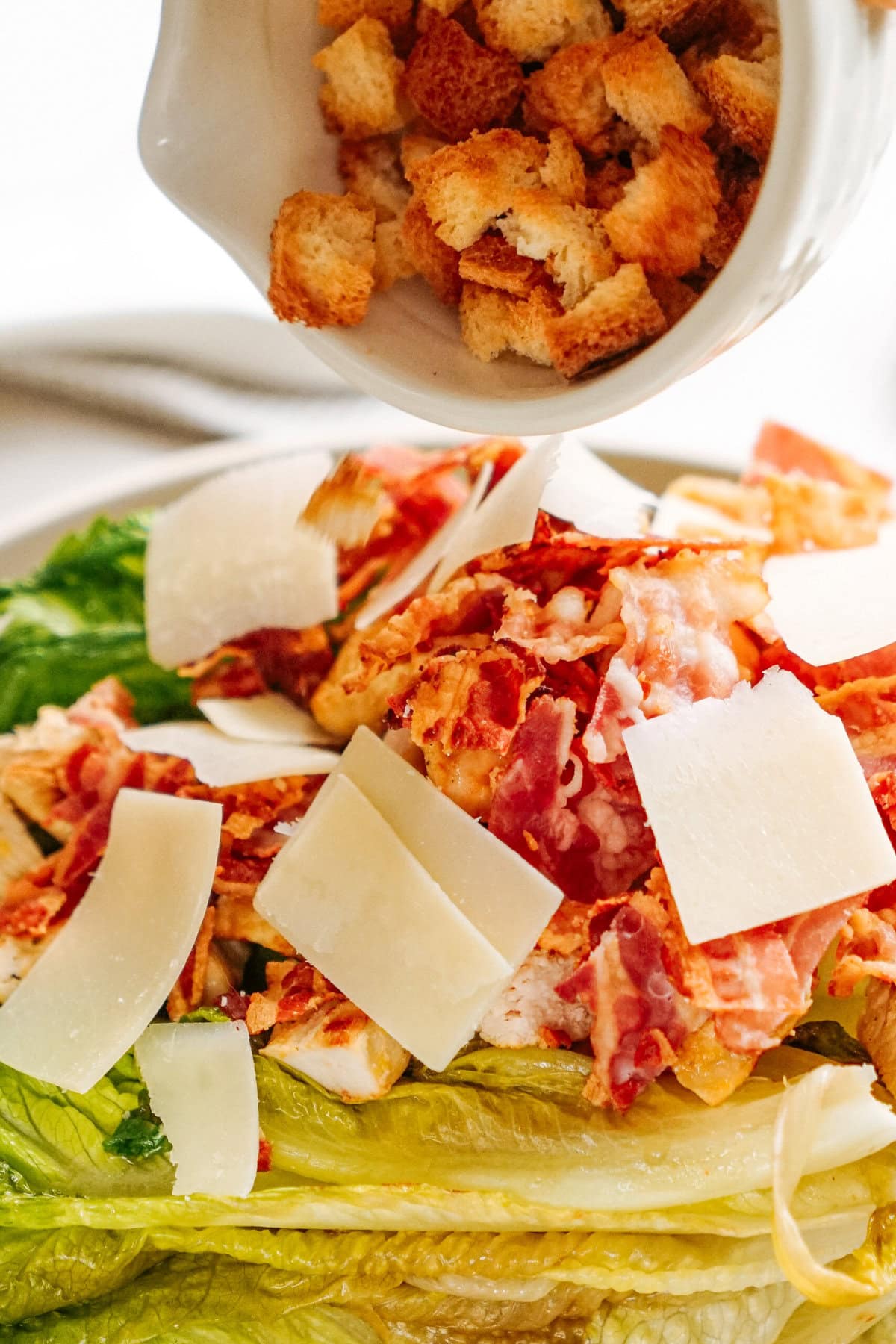 Croutons being added to a Caesar salad with lettuce, bacon, and cheese shavings.