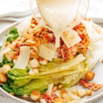 A grilled romaine salad topped with croutons, bacon, and thin cheese slices is drizzled with creamy dressing from a glass pitcher.