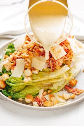 A grilled romaine salad topped with croutons, bacon, and thin cheese slices is drizzled with creamy dressing from a glass pitcher.