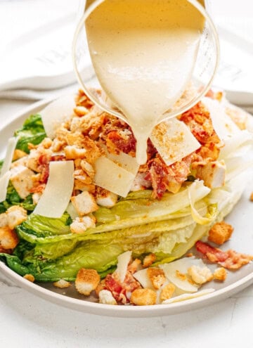 A grilled romaine salad topped with croutons, bacon, and thin cheese slices is drizzled with creamy dressing from a glass pitcher.