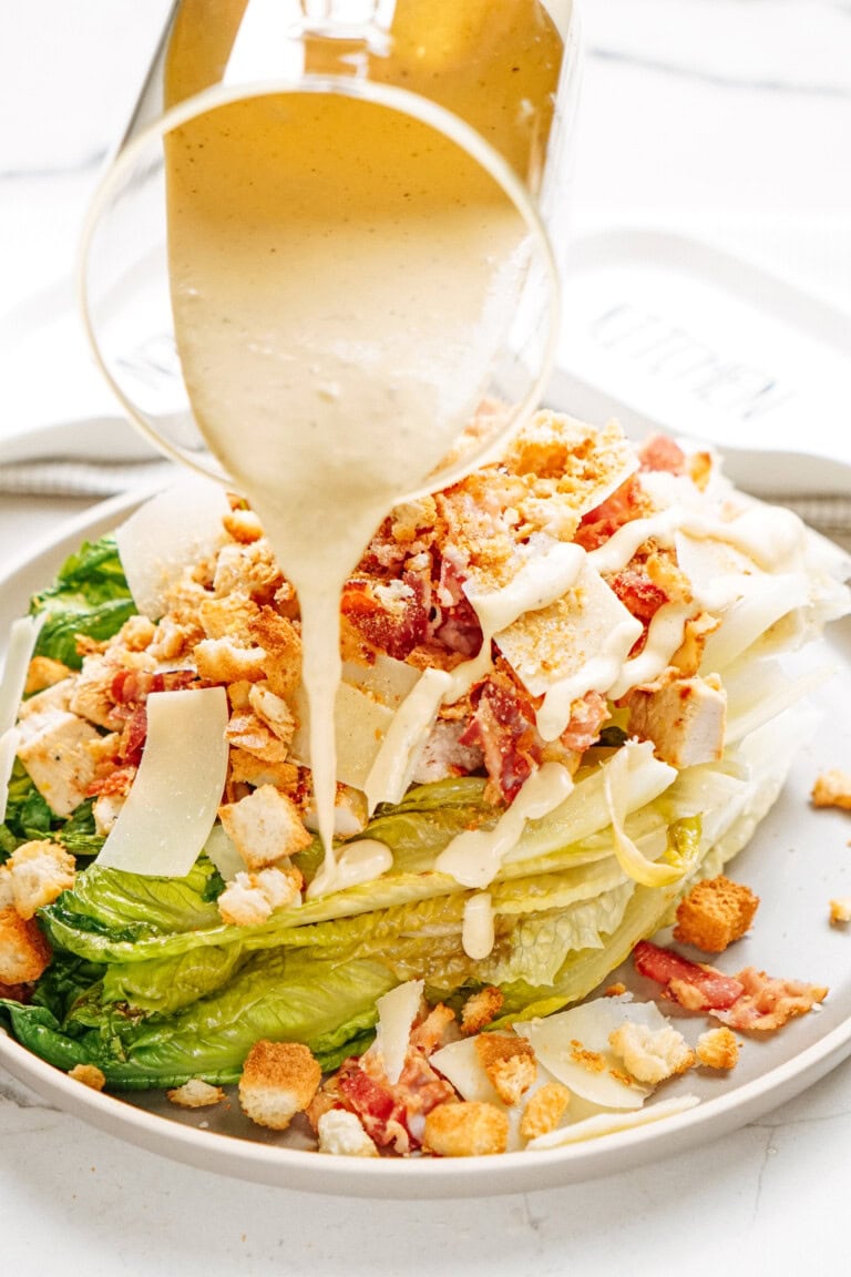 A grilled romaine salad topped with croutons, bacon, and thin cheese slices is drizzled with creamy dressing from a glass pitcher.