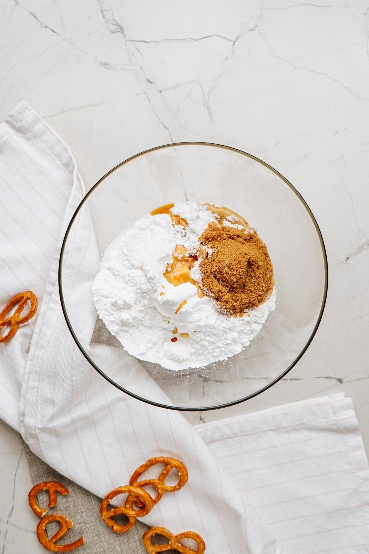 A glass bowl brimming with powdered sugar, brown sugar, and creamy peanut butter sits on a marble surface, embodying the essence of a Peanut Butter Cookie Dough Dip recipe. Scattered pretzels and a soft white cloth complete this inviting scene.