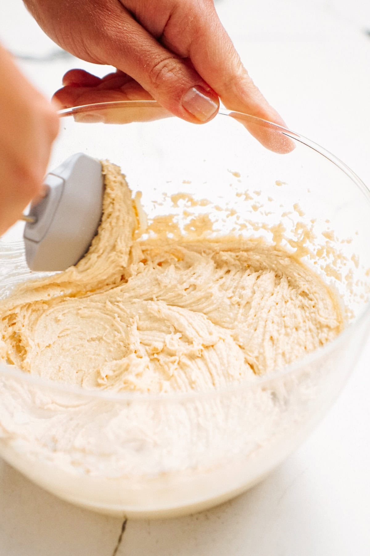 Hands mixing peanut butter cookie dough dip in a clear glass bowl with a spatula on a white surface.