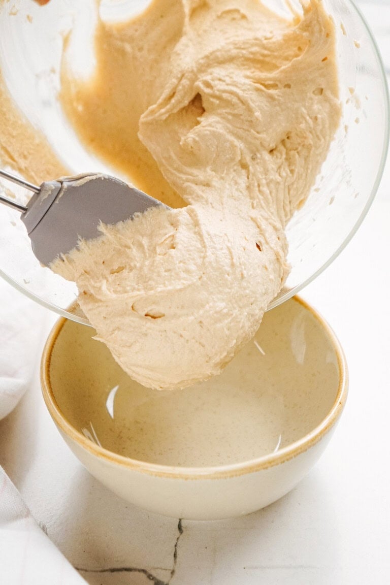 A mixer beater with creamy peanut butter cookie dough dip is poised above an empty beige bowl on a marble countertop.