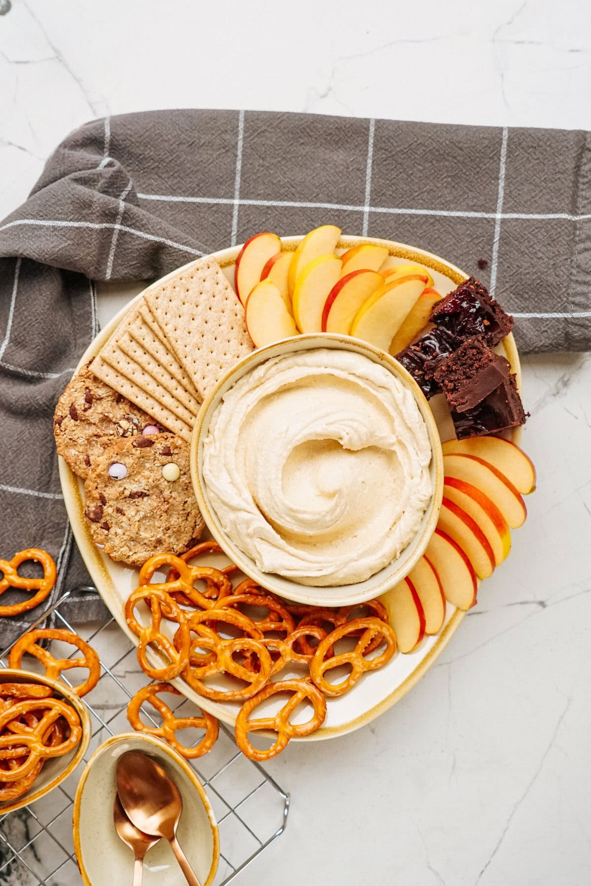 A platter featuring a bowl of hummus, surrounded by apple slices, crackers, and peanut butter cookie dough dip; complemented by cookies, brownies, and pretzels on a checkered cloth backdrop.