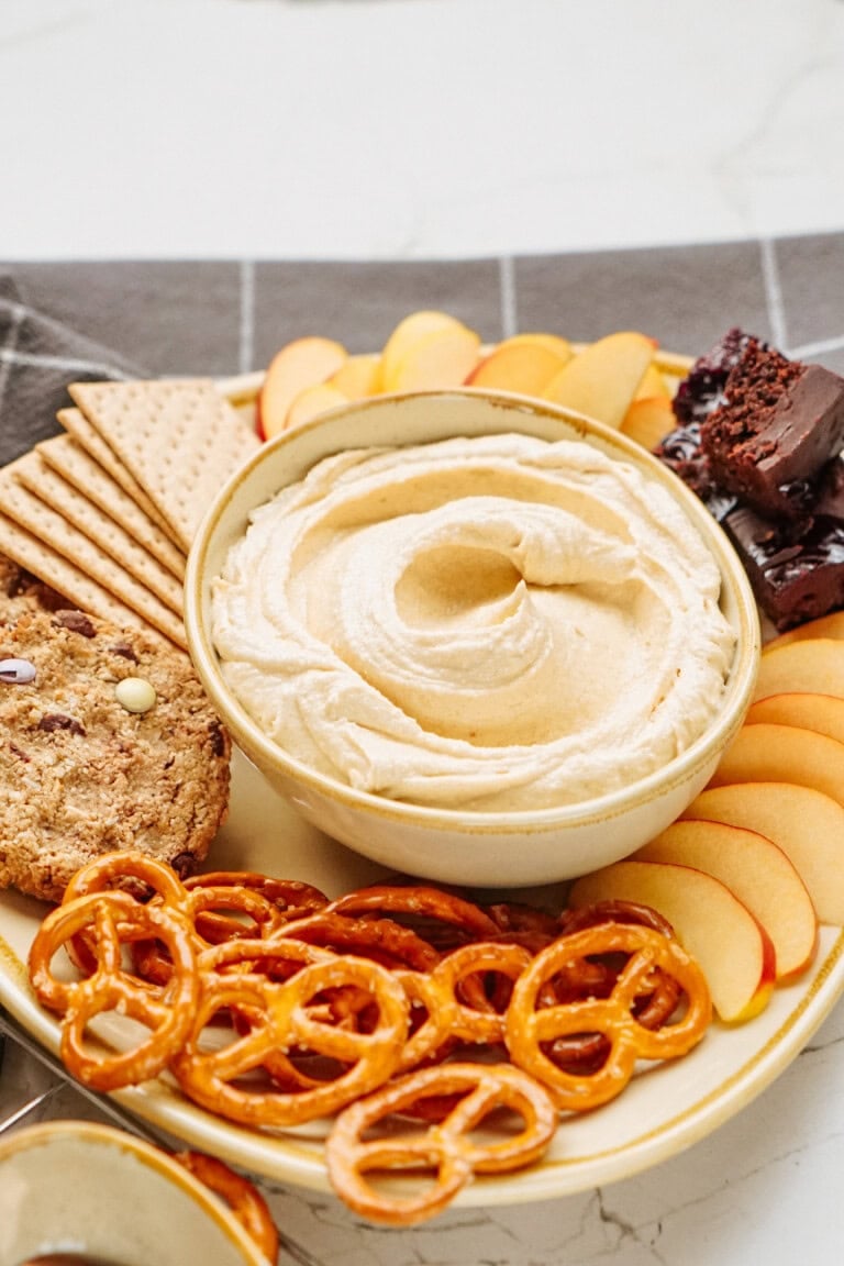 A platter with a bowl of delicious peanut butter cookie dough dip, accompanied by sliced apples, pretzels, crackers, and cookies, all artfully arranged for serving.