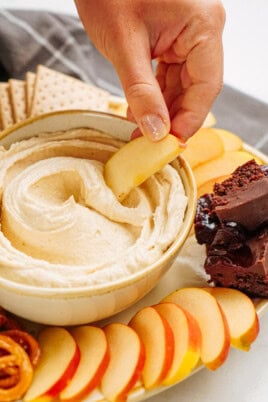 A hand dips an apple slice into a bowl of creamy peanut butter cookie dough dip on a platter with apple slices, chocolate brownies, pretzels, and crackers.