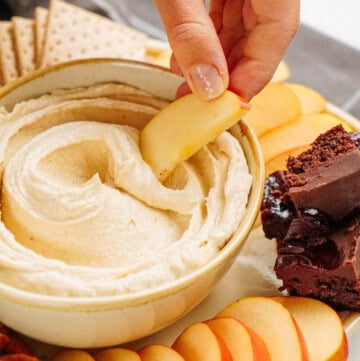 A hand dips an apple slice into a bowl of creamy peanut butter cookie dough dip on a platter with apple slices, chocolate brownies, pretzels, and crackers.