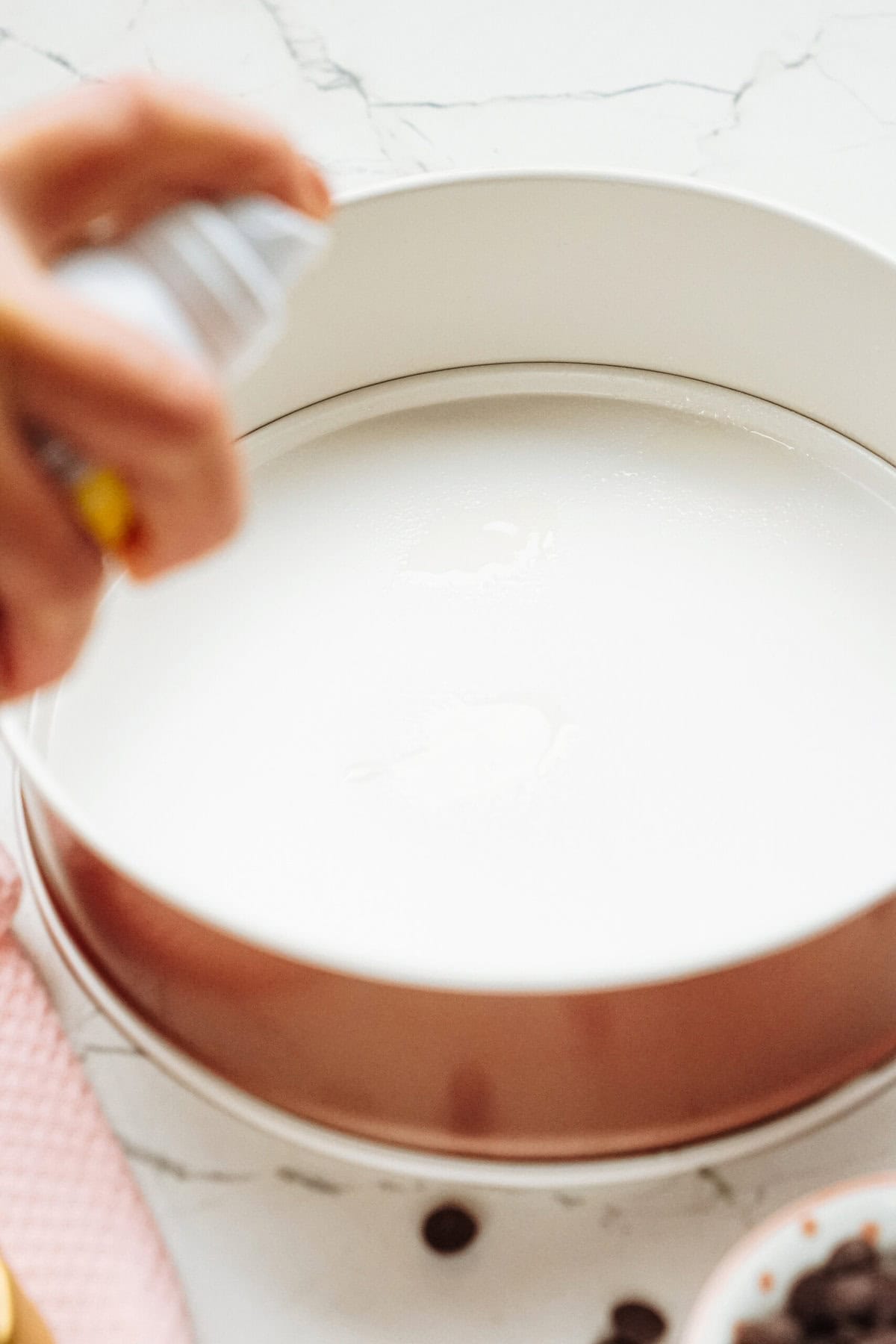 A hand sprays non-stick spray into a round baking pan on a marble countertop.