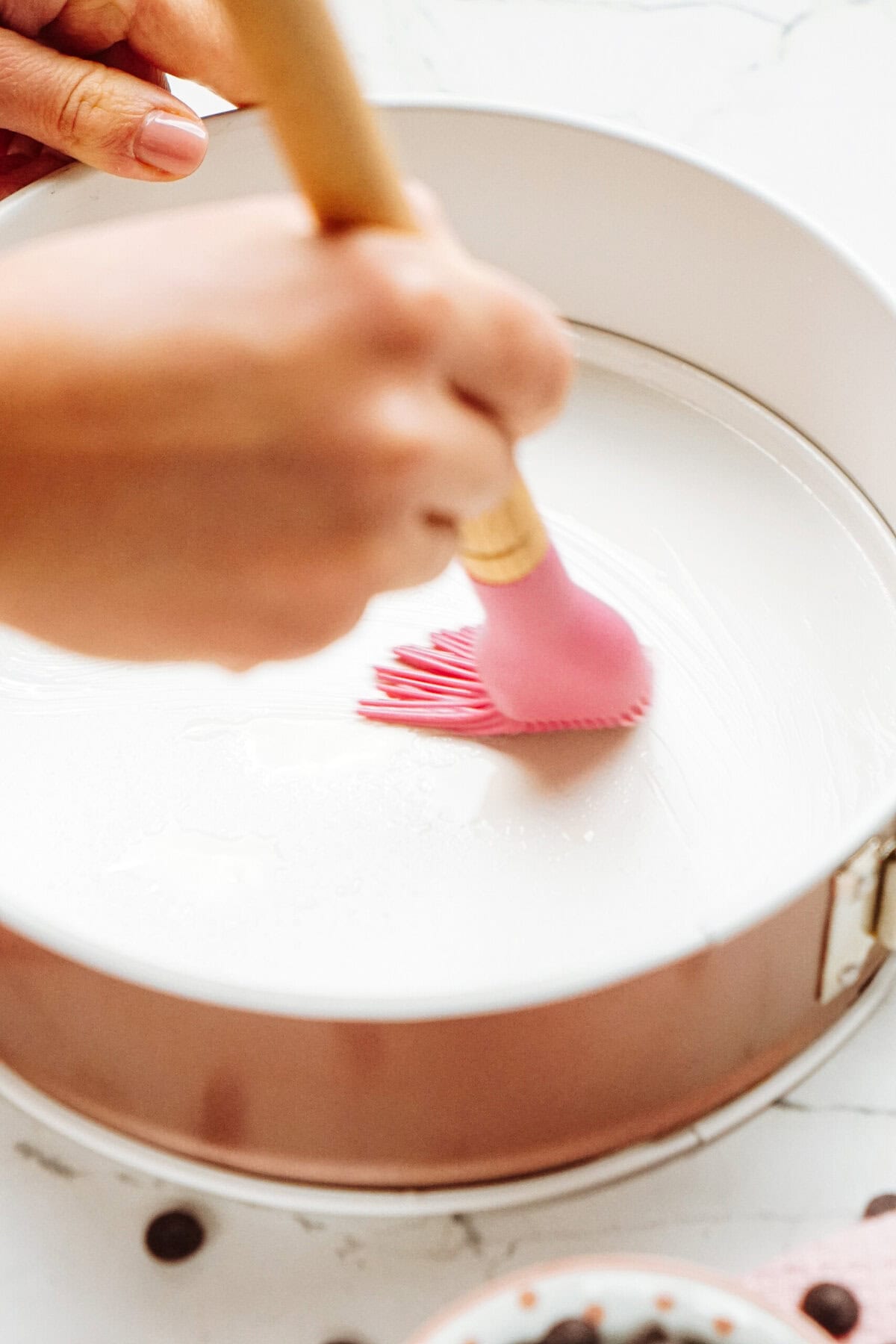 A person uses a pink silicone brush to spread white substance inside a round cake pan on a white surface.