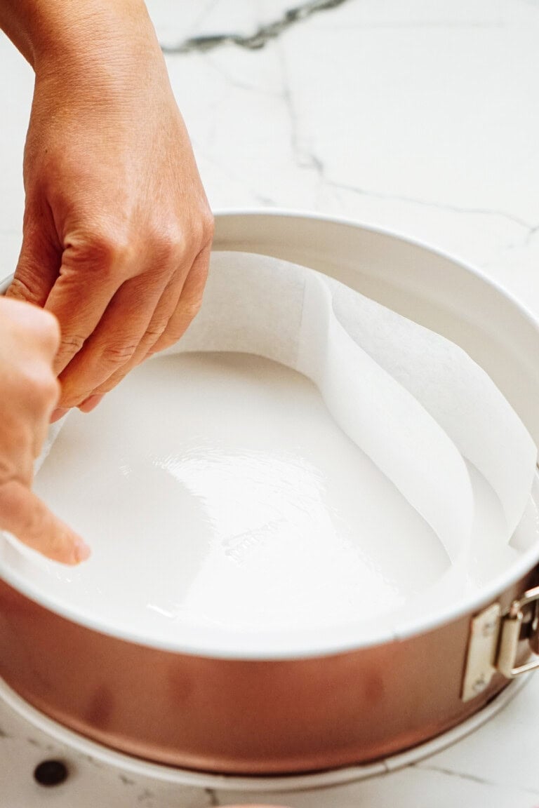 Hands lining a round cake pan with parchment paper on a marble surface.