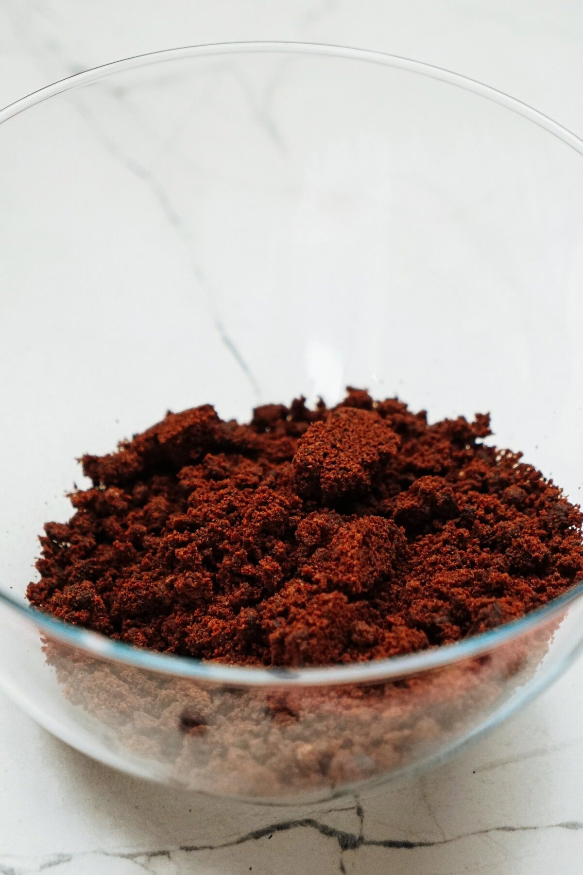 A glass bowl containing a pile of ground coffee on a marble surface.