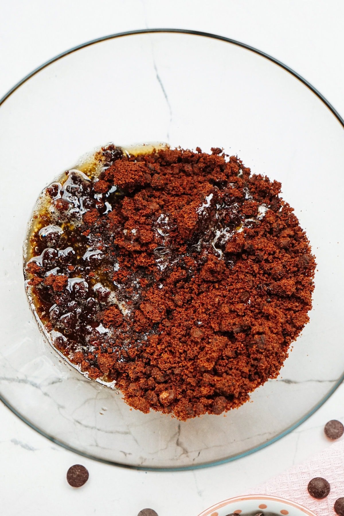 A round chocolate pudding with a thick crumbly topping sits on a glass plate, placed on a white marble surface.