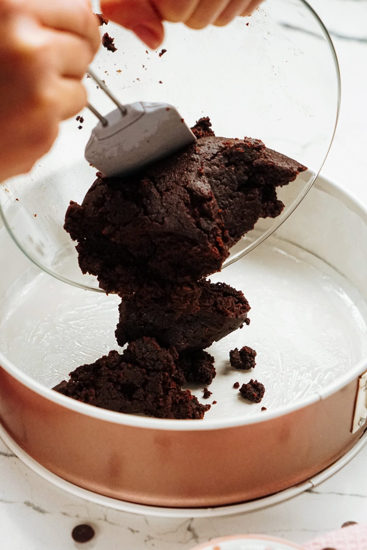 Hands transferring thick, dark chocolate batter from a glass bowl into a round baking pan with a spatula.