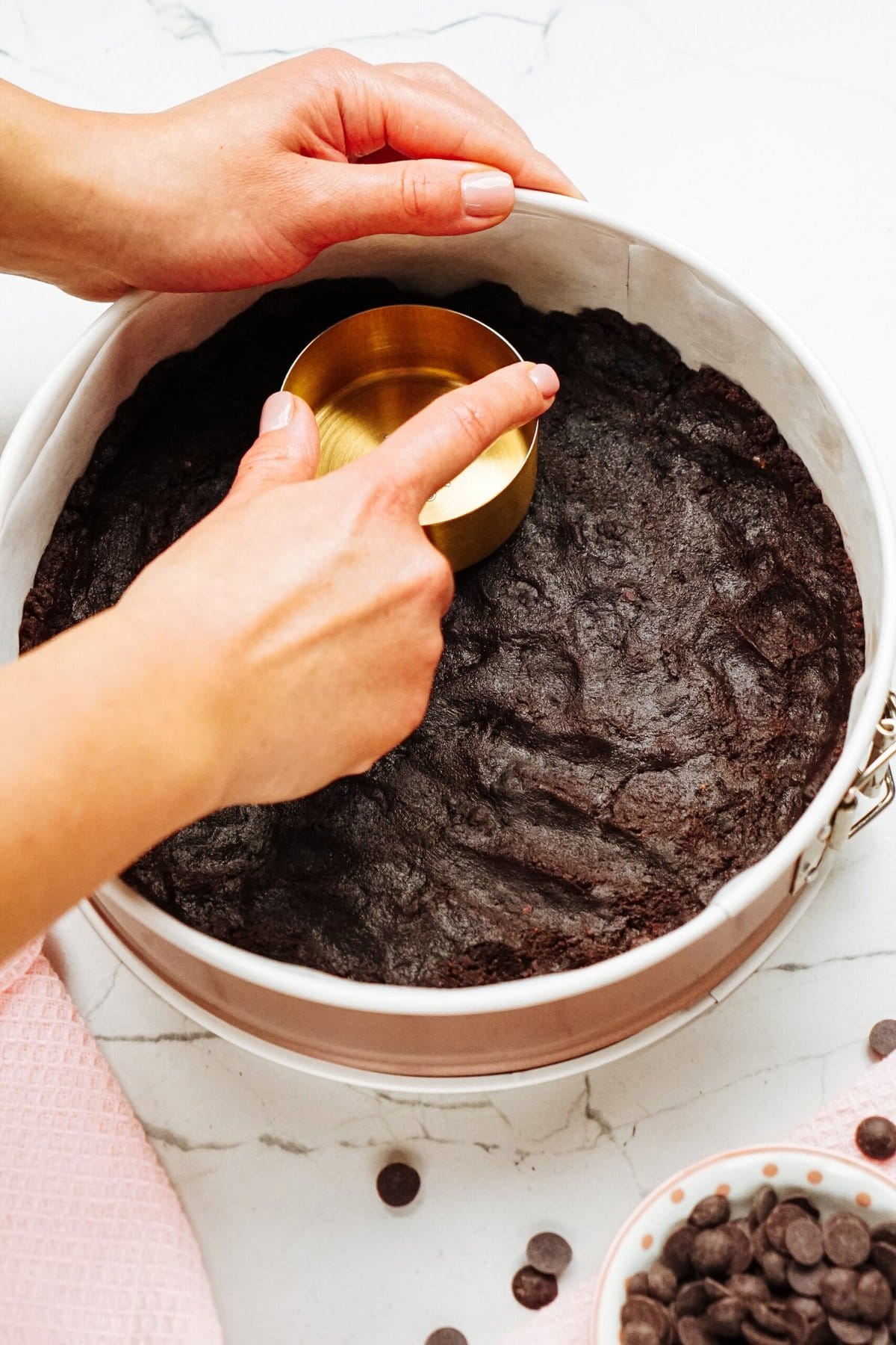 Hands pressing chocolate crust into a round pan with a measuring cup, on a marble countertop. A bowl of chocolate chips is nearby.