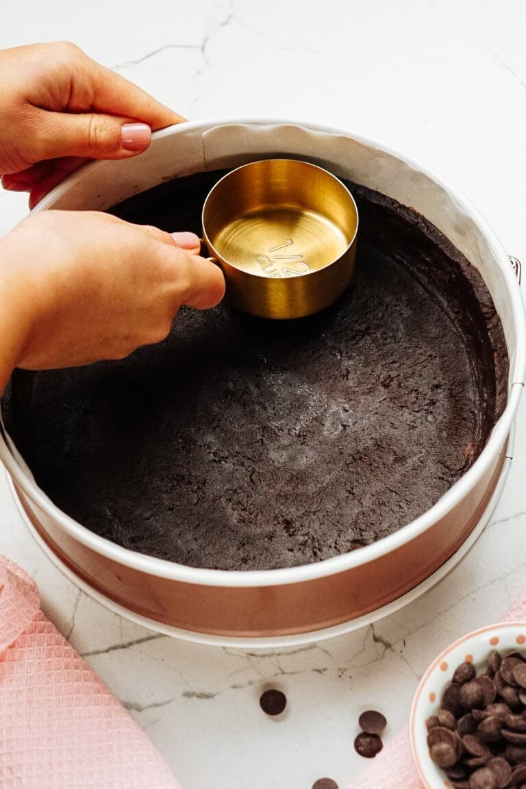 Hands pressing crumbs into a round pan with a measuring cup on a marble surface. Nearby are a pink cloth and a bowl of chocolate chips.