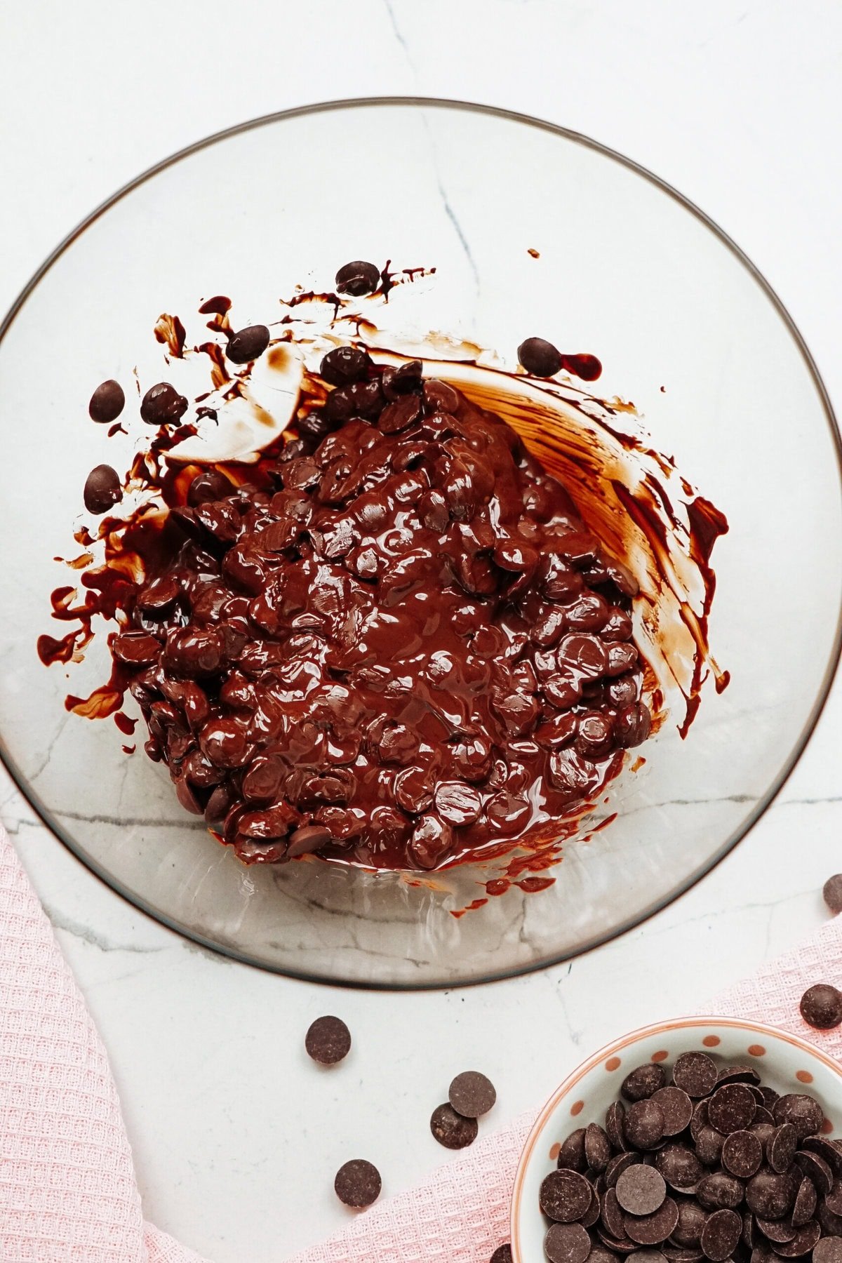 A glass bowl filled with a mixture of melted chocolate and chocolate chips. There's some chocolate smeared on the side of the bowl. A small bowl of chocolate chips is nearby, with a pink cloth.