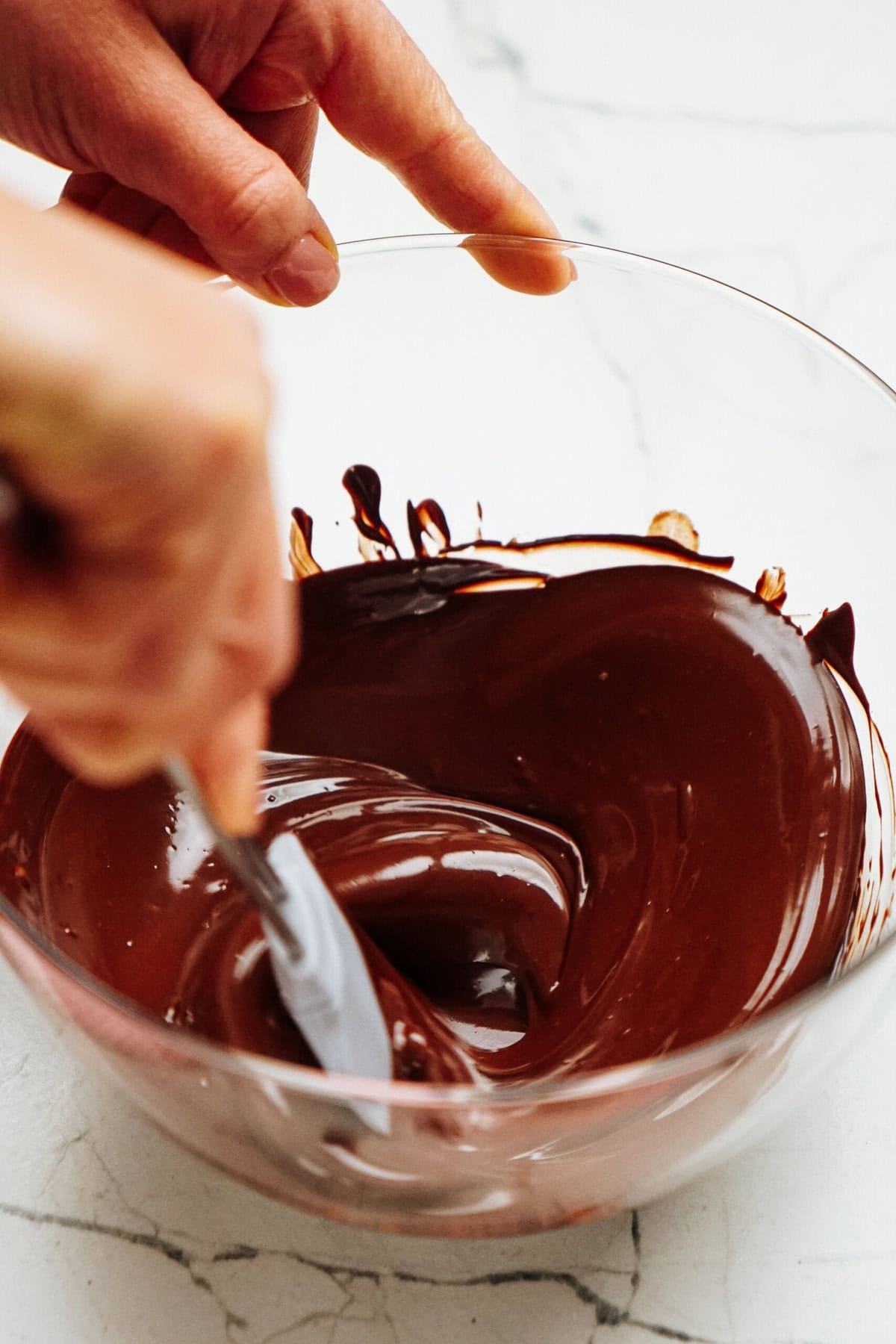 A person stirring melted chocolate in a clear glass bowl with a spatula on a marble countertop.