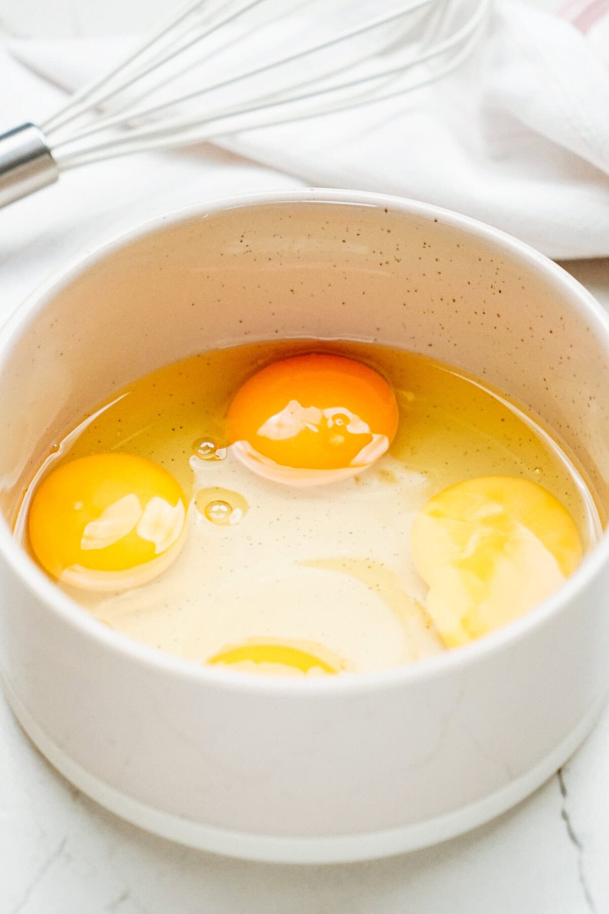 Three raw eggs in a white bowl with a whisk in the background on a white surface.