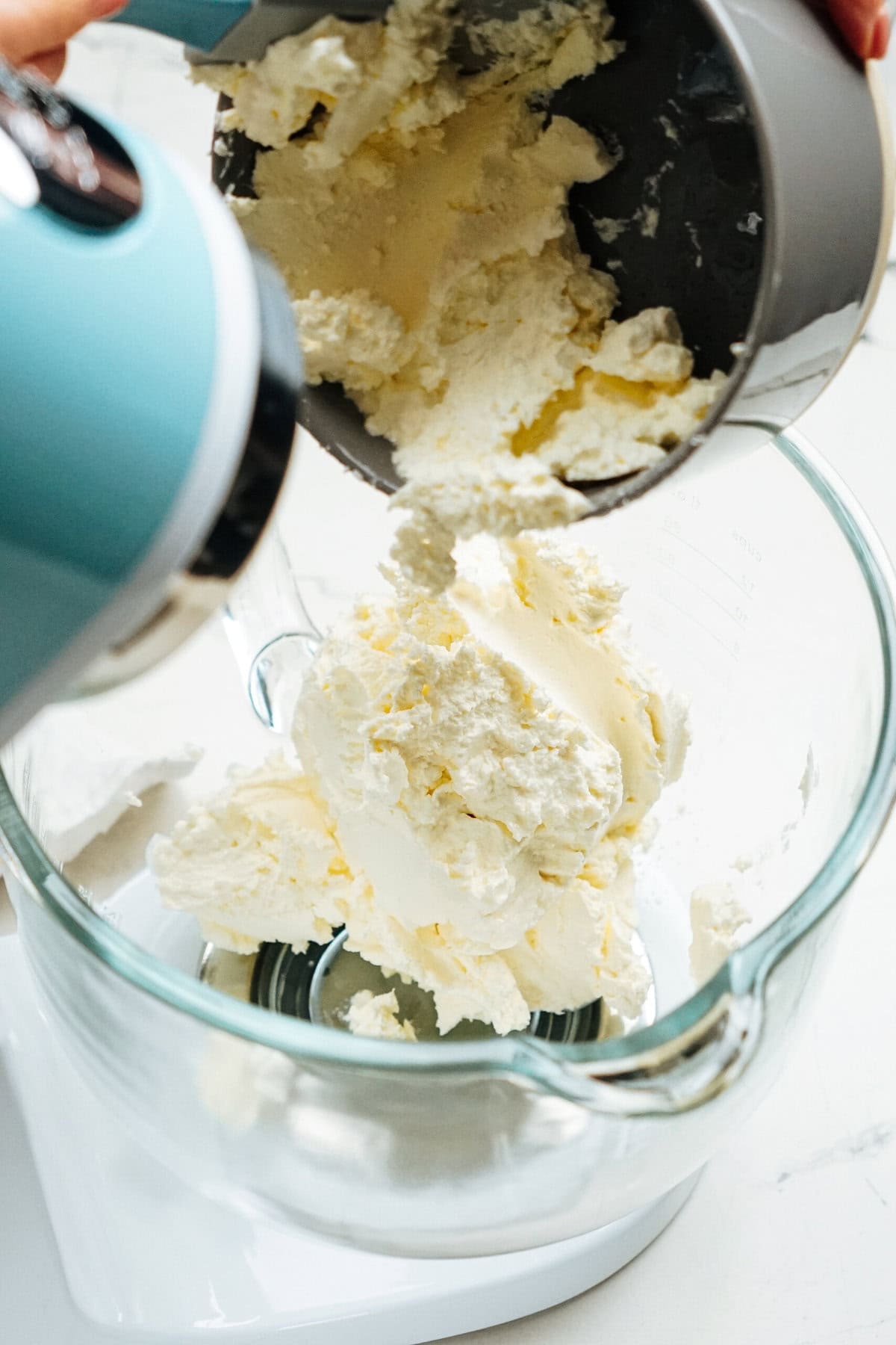 Cream is being transferred from a mixing bowl to a glass stand mixer bowl.