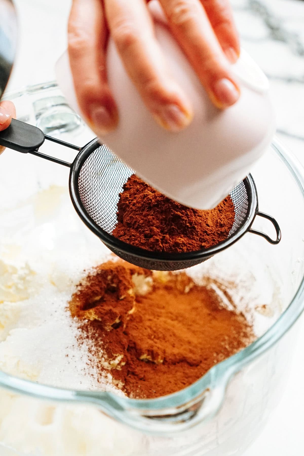 Person sifting cocoa powder and spices into a bowl containing other ingredients.