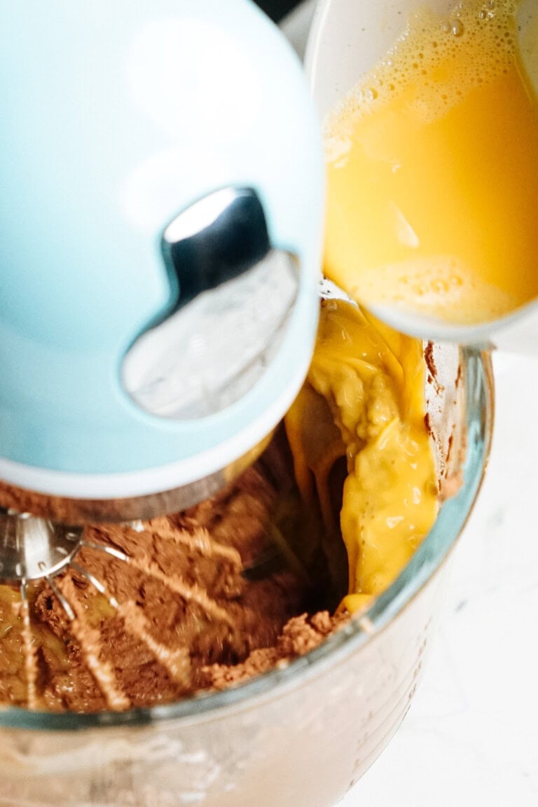 A blue stand mixer blending chocolate batter as beaten eggs are being poured into the bowl.