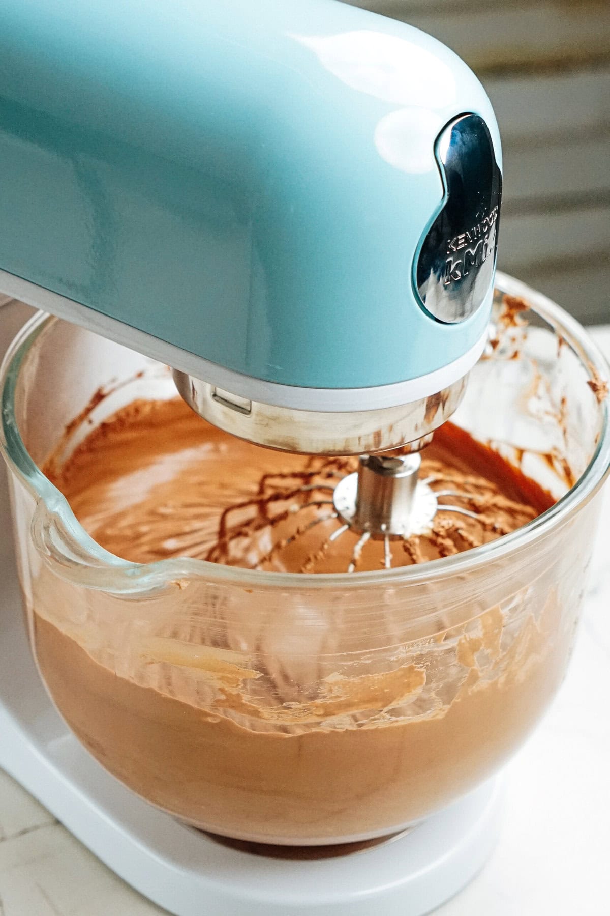 A light blue stand mixer with a glass bowl is mixing chocolate batter with a wire whisk attachment.