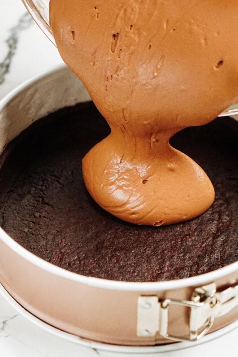 Pouring chocolate batter over a baked base in a springform pan on a marble surface.