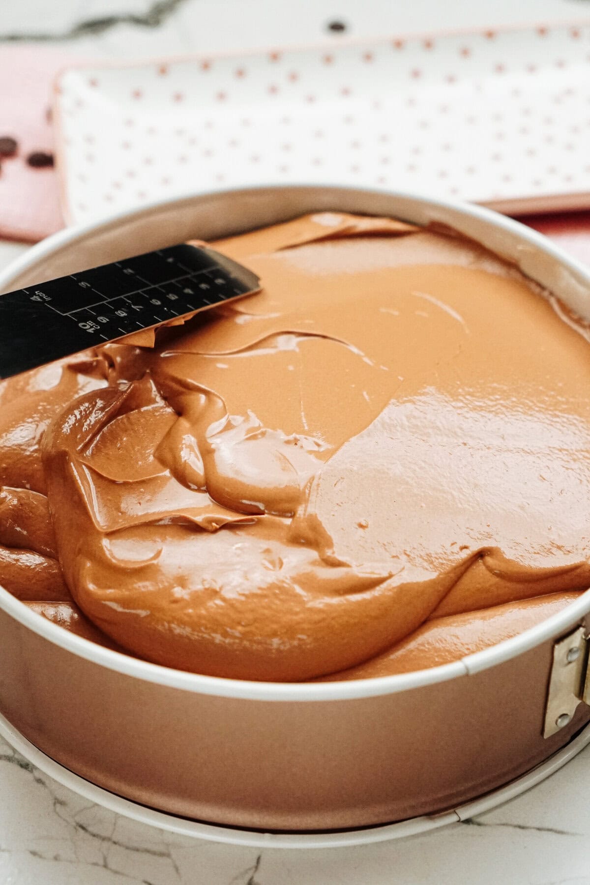 Smooth cheesecake layer in a round baking pan with a metal spatula on top.