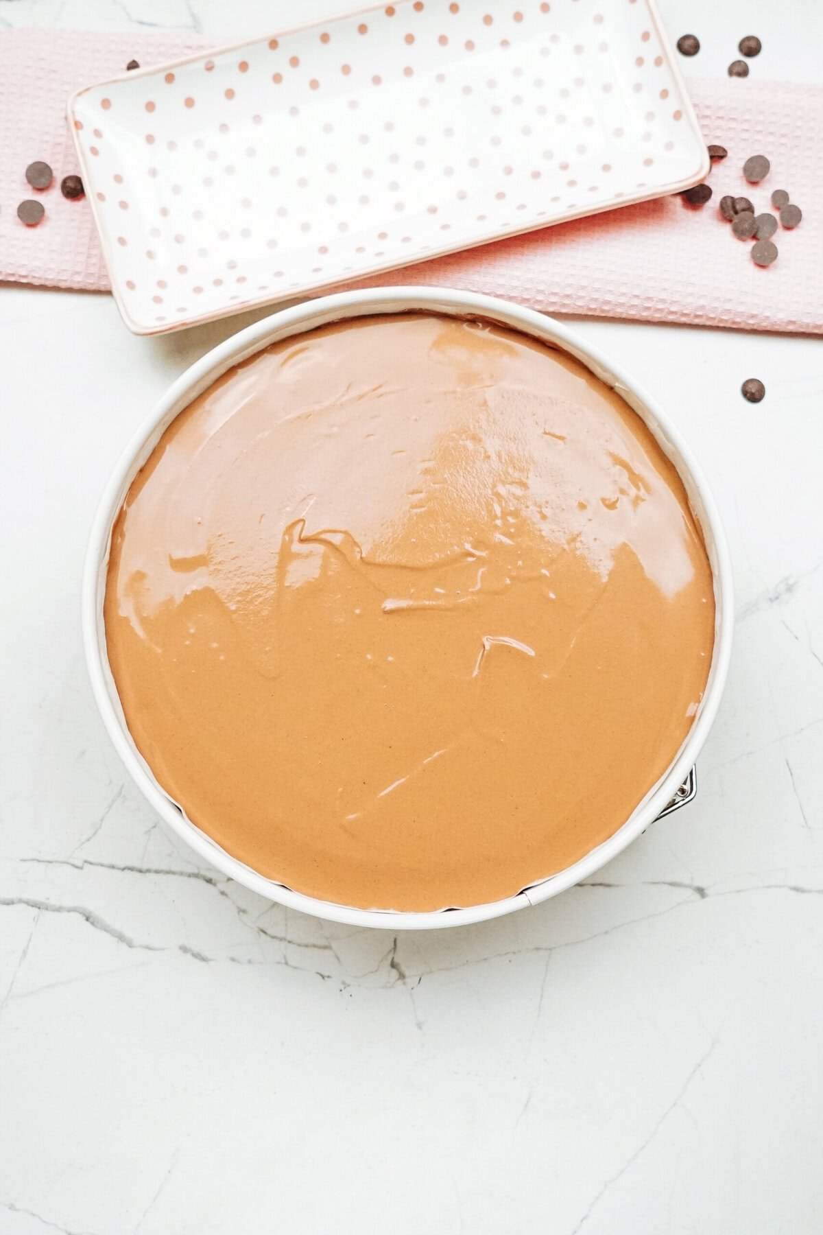 A round bowl of smooth light brown batter sits on a marble surface. A rectangular dish with a dotted pattern and scattered chocolate chips is in the background.