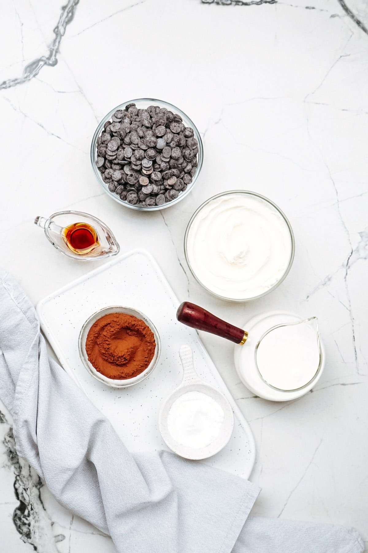 Ingredients for a delightful chocolate mousse await on the marble countertop: chocolate chips, vanilla extract, whipped cream, milk, cocoa powder, and a mysterious white powder in dishes. A blue cloth is draped nearby.