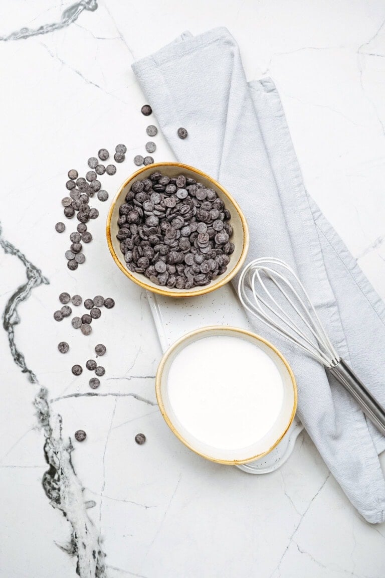 Bowl of chocolate chips and bowl of milk on a marble surface, with a whisk and a gray cloth nearby.