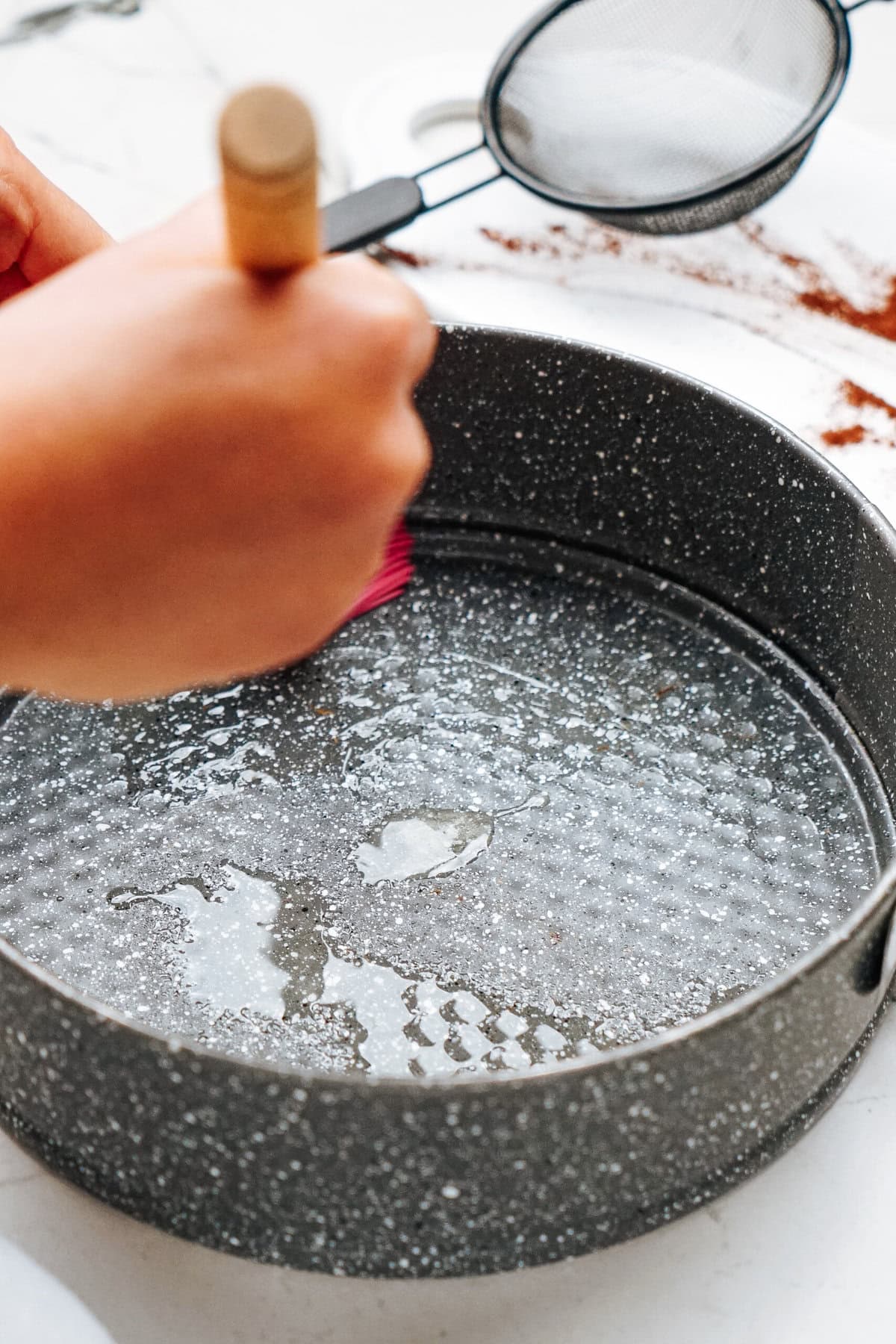 Hands greasing a round baking pan with a brush; a sieve and some spilled flour are visible in the background.