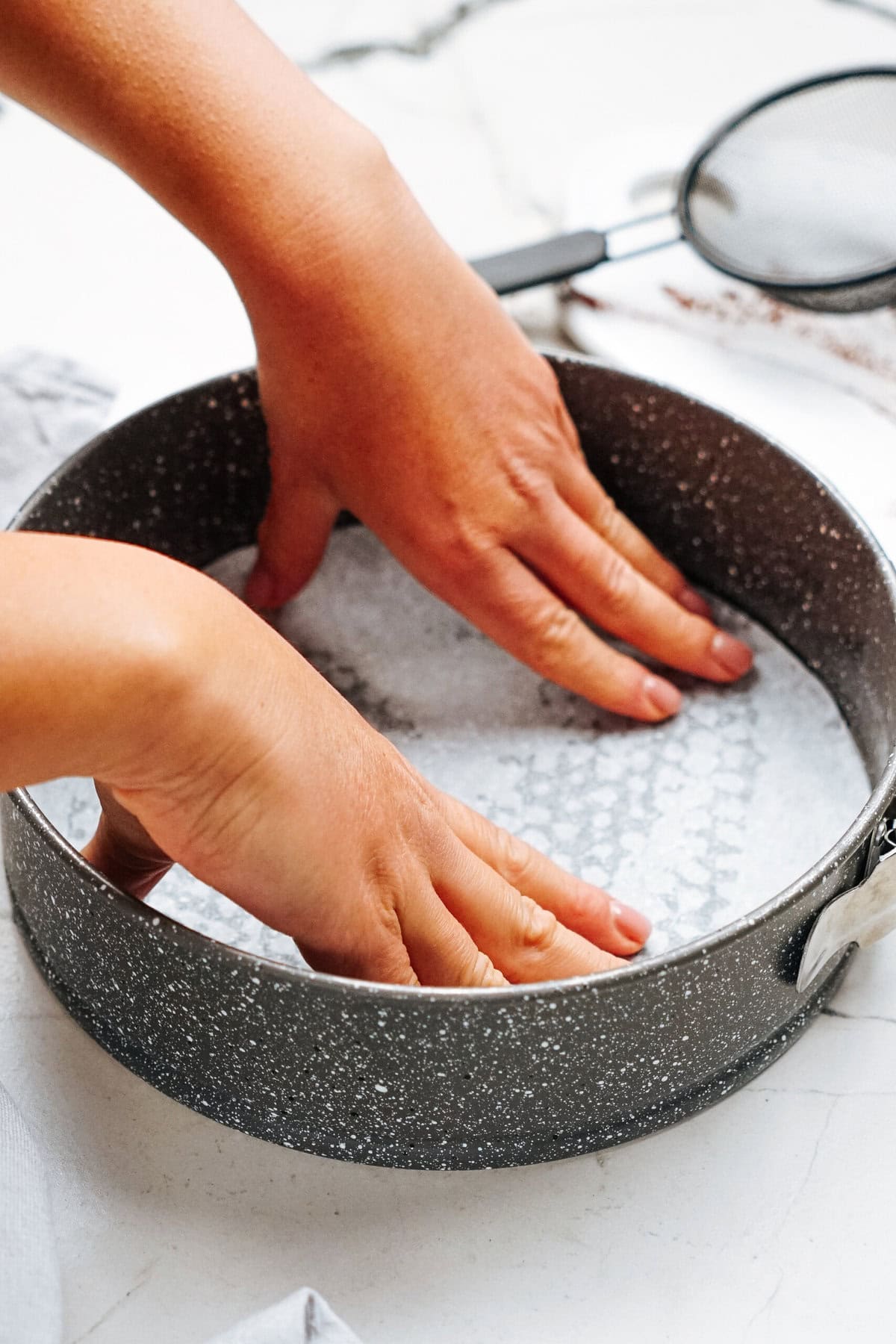 Hands pressing parchment paper into a round baking pan on a marble surface. A sifter is nearby.