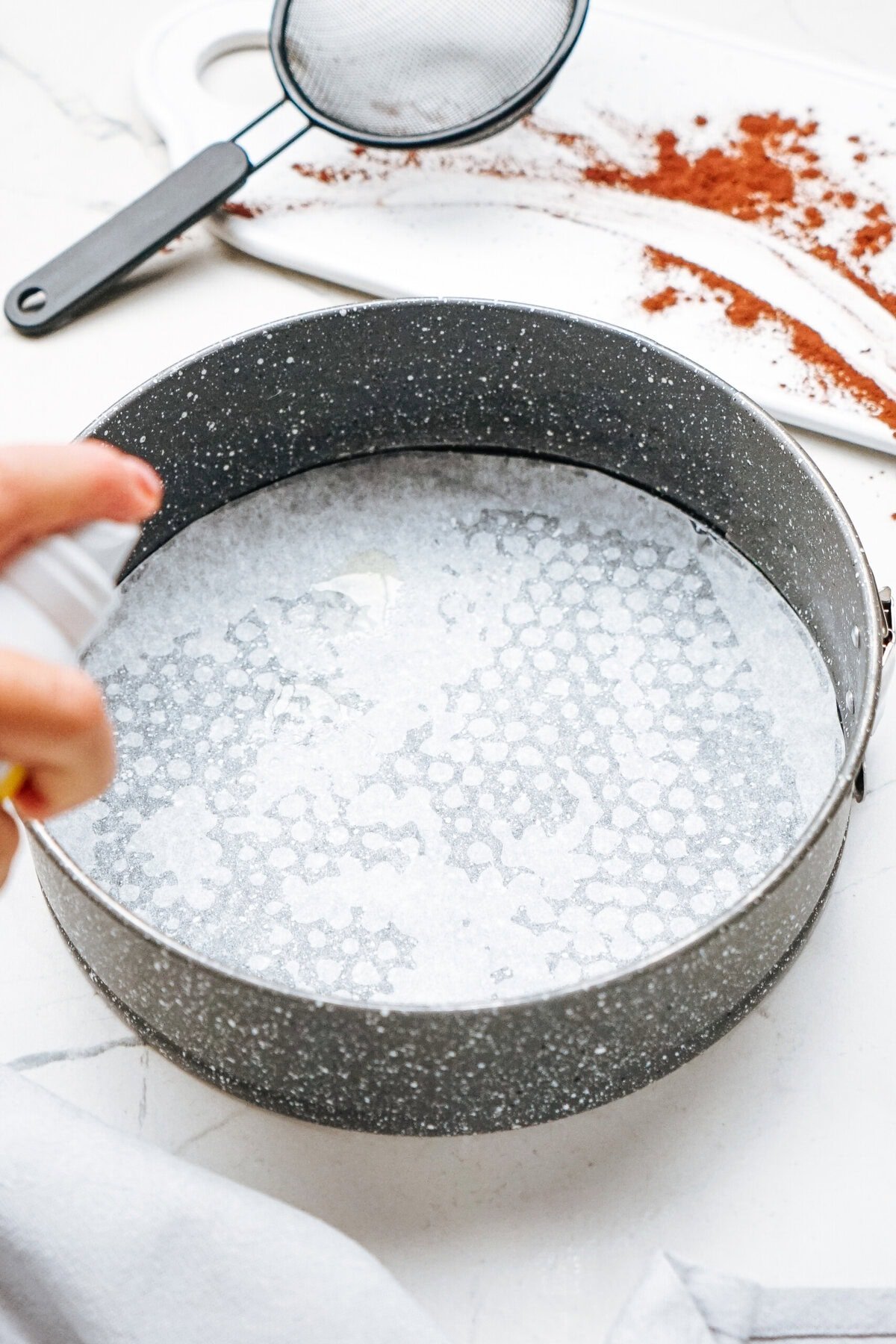 A round baking pan with parchment paper, being sprayed with cooking spray. A sieve and cocoa powder are on a white board in the background.