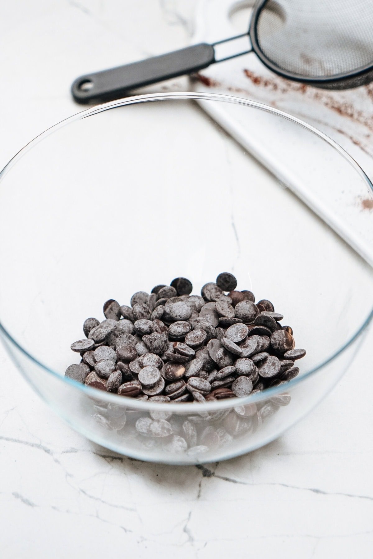 A glass bowl filled with chocolate chips on a marble countertop, with a sifter and a cutting board in the background.