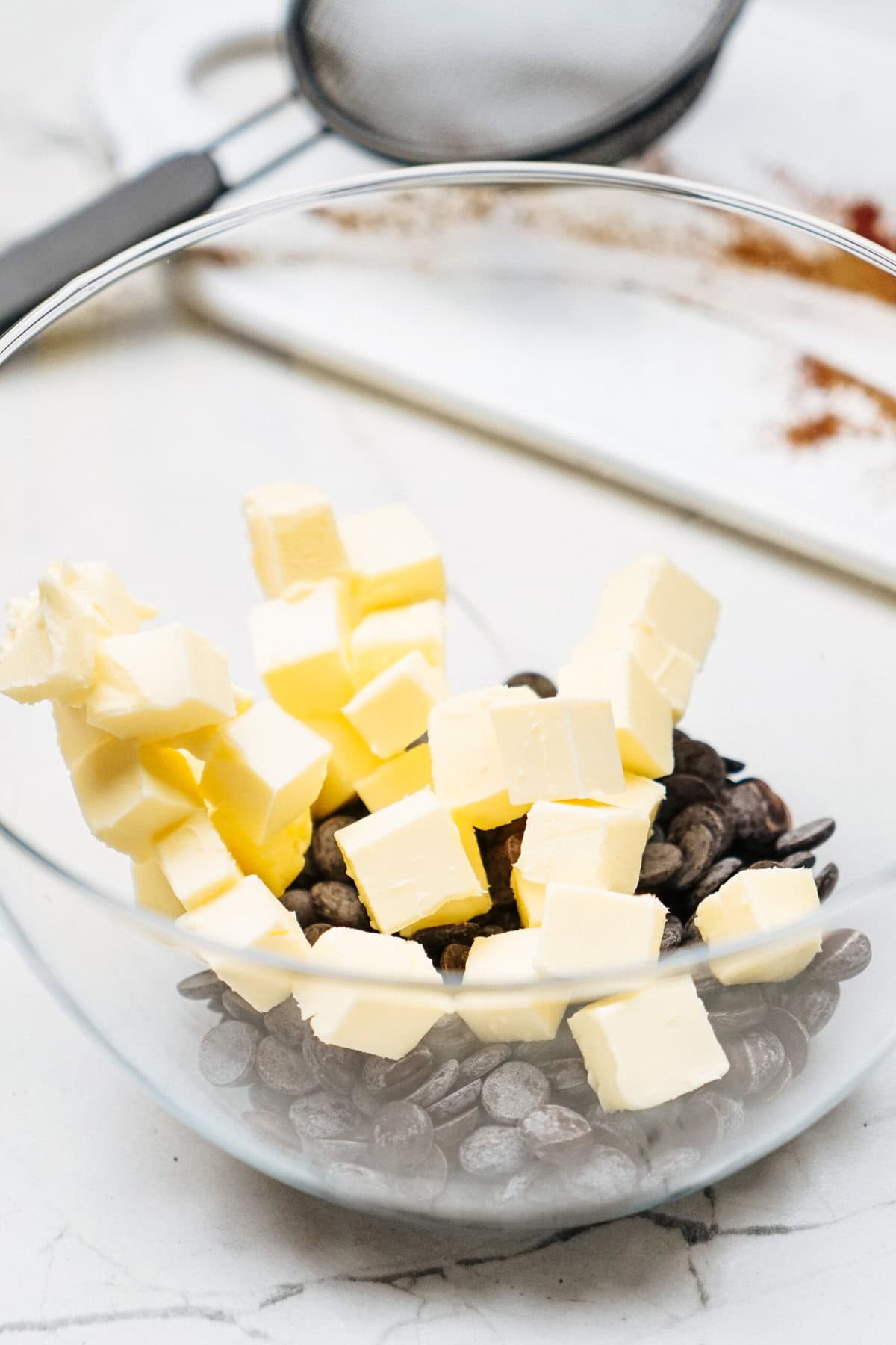 Glass bowl with cubes of butter and chocolate chips on a marble countertop, with a strainer and cutting board in the background.