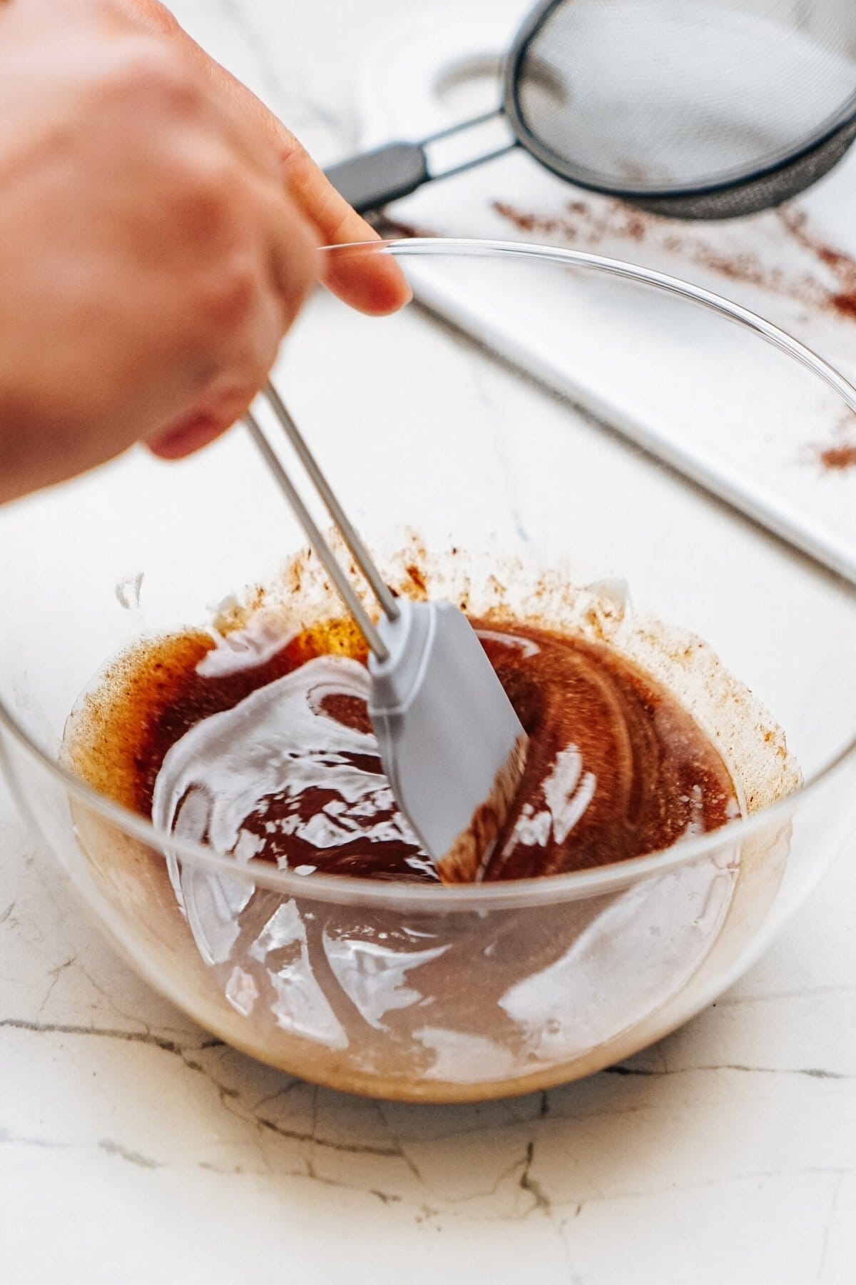 Person mixing brown liquid in a clear glass bowl with a spatula.