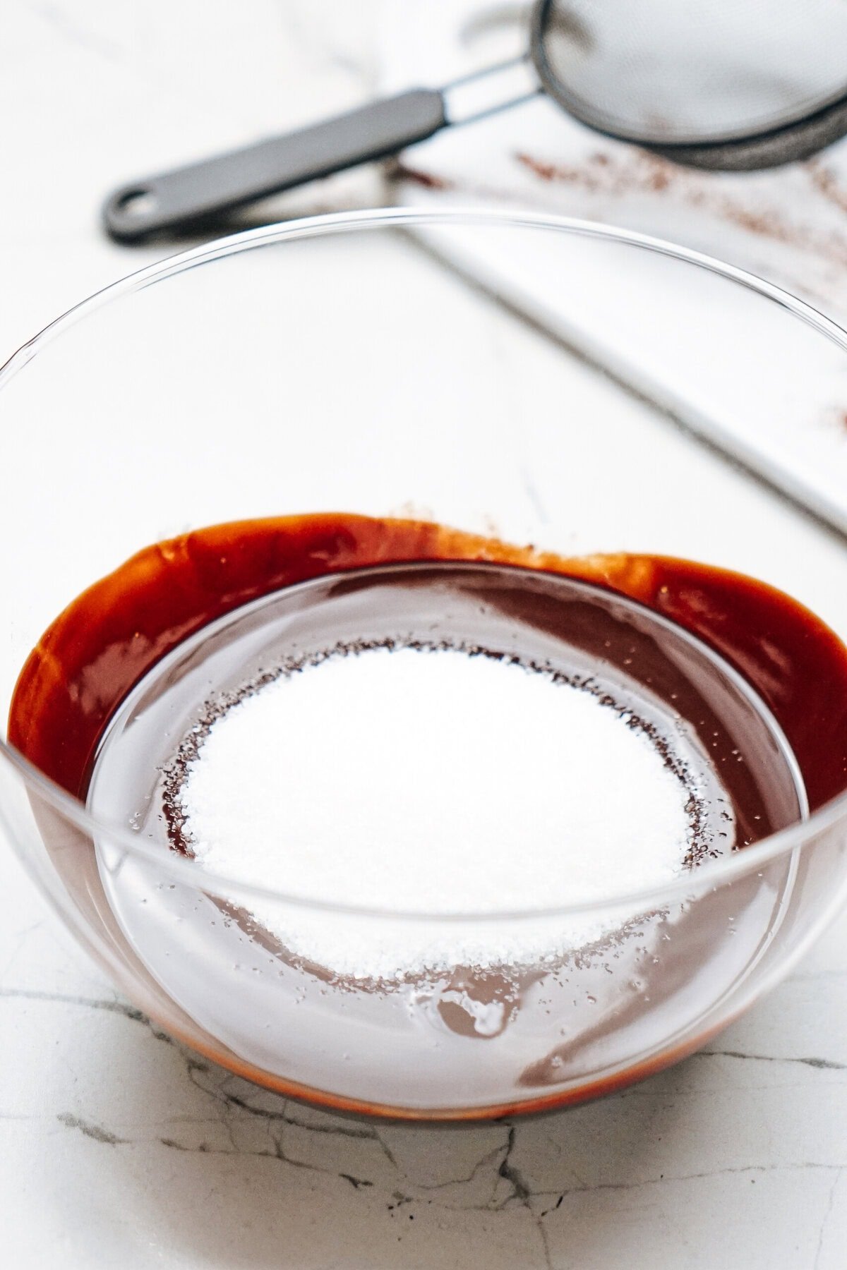 A clear bowl containing chocolate liquid with a mound of white sugar on top, placed on a white countertop near a metal strainer and a cloth.