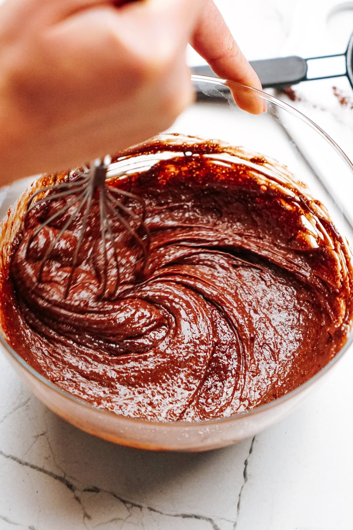 A hand whisking chocolate cake batter in a glass bowl on a marble countertop.