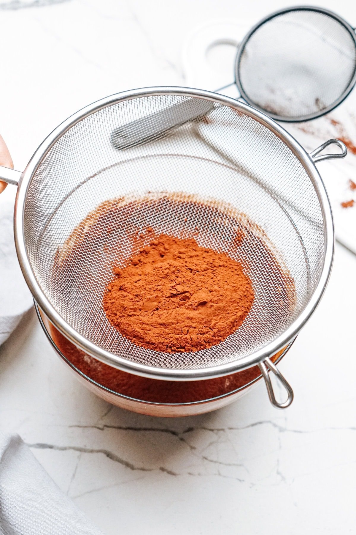 A metal sieve containing cocoa powder placed over a bowl on a marble countertop.