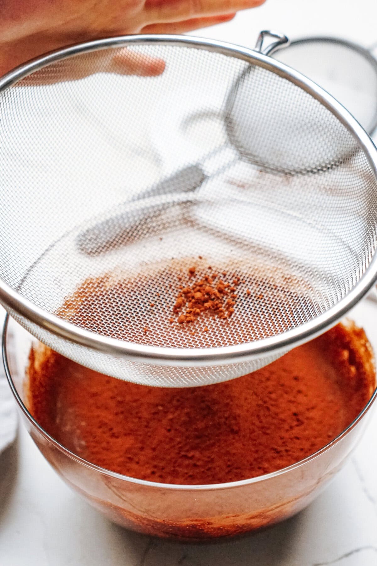 A hand sifting cocoa powder into a glass bowl using a metal sieve on a white surface.