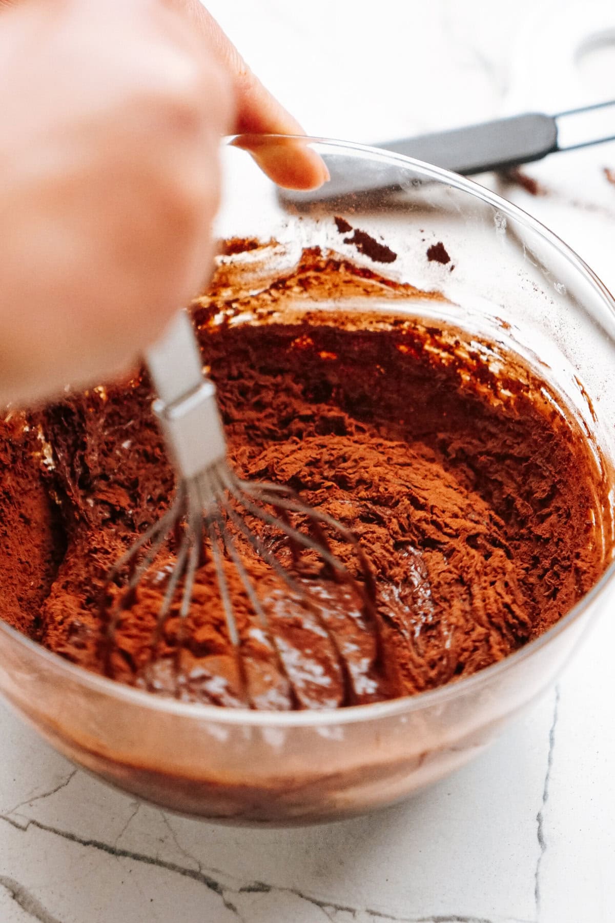 A hand whisks cocoa powder mixture in a clear glass bowl on a marble countertop.