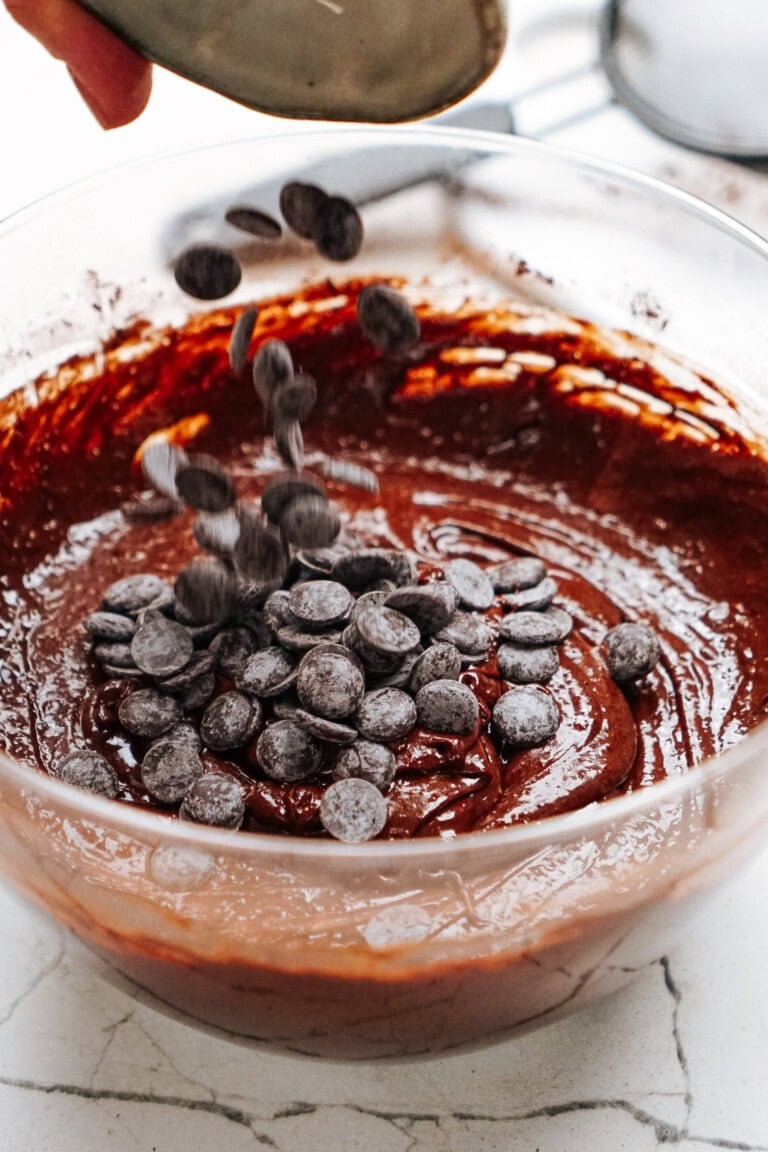 A bowl of chocolate batter with chocolate chips being poured into it on a marble countertop.